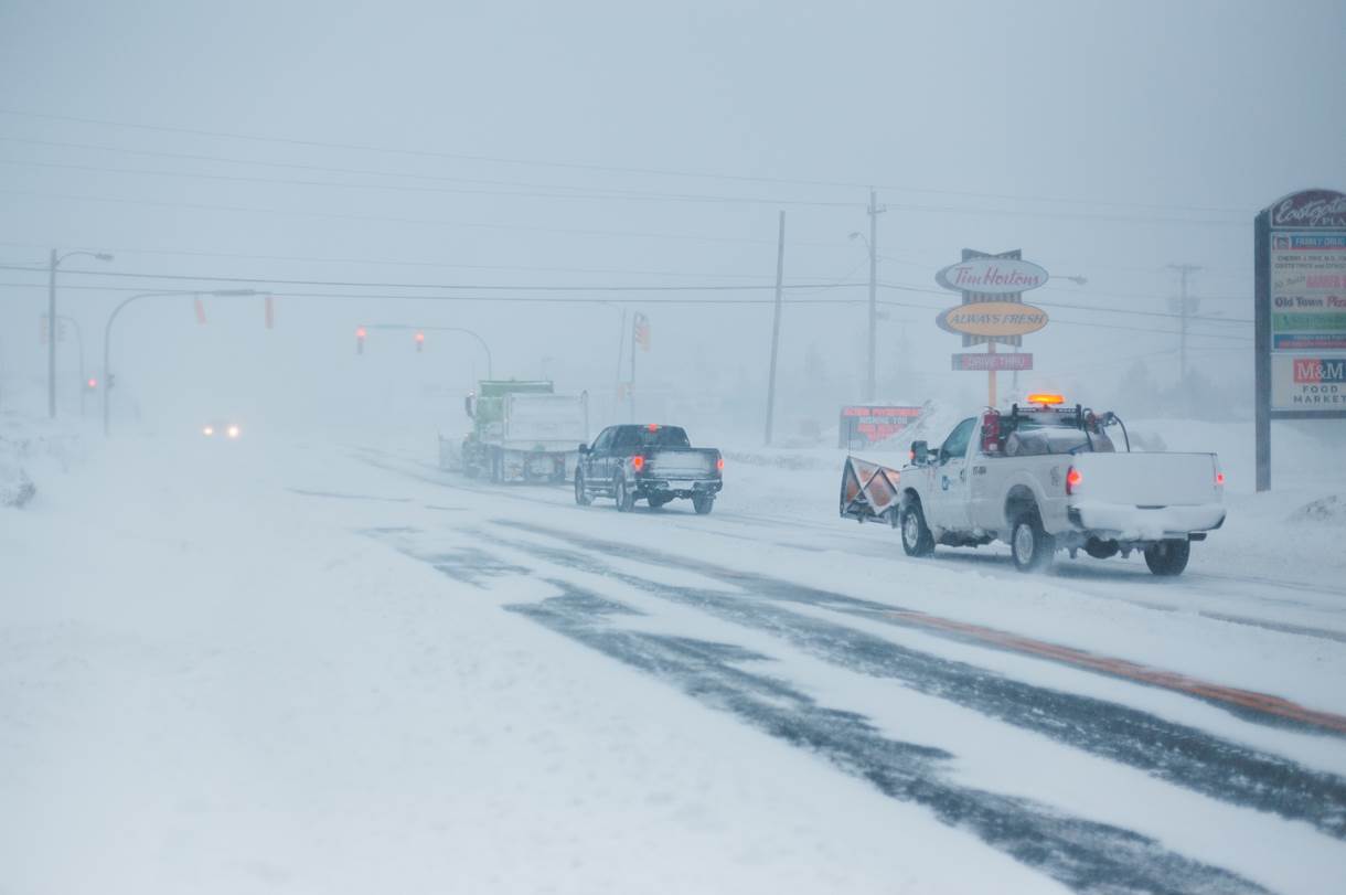 Snowplows and vehicles navigate a snow-covered road, battling heavy snowfall. Signs for "Tim Hortons" and "Old Town Plaza" are visible amidst the low visibility, illustrating challenging transport conditions.