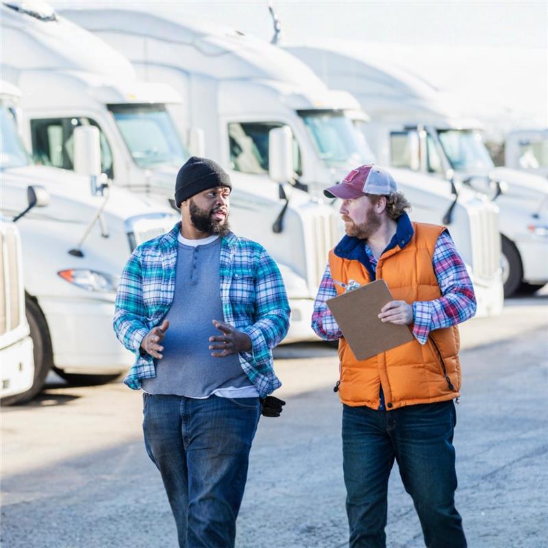 Two men stand in front of a truck, discussing a clipboard. One wears a plaid shirt, looking thoughtful, while the other, in work attire and gloves, explains details.